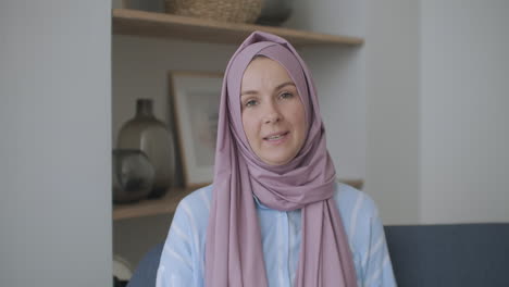Looking-at-the-camera-muslim-girl-calling-friends-from-apartments-using-a-video-link-waving-with-hand-sitting-on-a-sofa-in-a-stylish-room-in-front-of-computer-screen-having-video-conversation